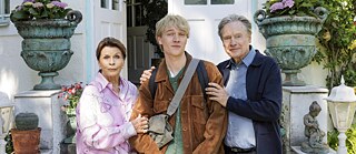 An older woman and an older man stand to the left and the right of a young man an hold him caringly by his arms.