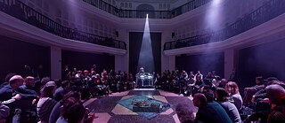 Audience seated in a circle inside a lit hall around a central performance and sound installation.