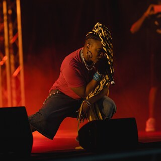 Personne tenant un micro, à genoux sur une scène de concert éclairée par des lumières rouges et envahie par la fumée.