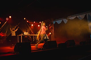 Une personne munie d'un micro se déplace sur la scène d'un concert. À l'arrière-plan, on aperçoit d'autres artistes, tandis que des projecteurs rouges et orange ainsi que de la fumée éclairent la scène.