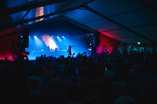 Vue sur le public, avec en arrière-plan la scène d'un concert dans un grand chapiteau. Des lumières bleues et rouges ainsi que des effets de fumée créent l'ambiance.