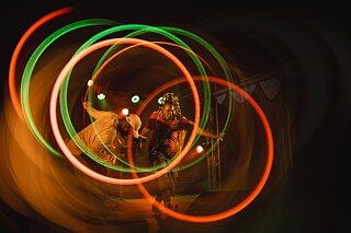 Une personne sur la scène d'un concert, entourée de traînées lumineuses rouges et vertes disposées en cercle, créées par une exposition longue. Une autre personne est visible à l'arrière-plan.