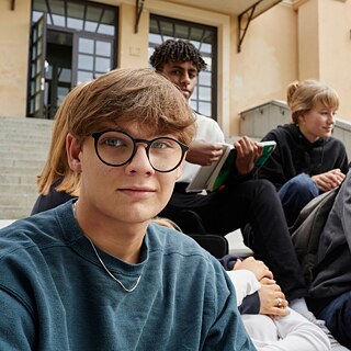 Eine Gruppe von Schüler*innen sitzt auf den Stufen vor ihrer Schule und wartet darauf, dass sie zur Deutschprüfung reingehen können.