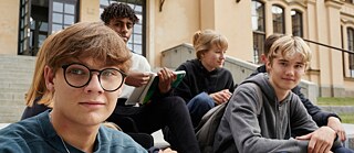 Eine Gruppe von Schüler*innen sitzt auf den Stufen vor ihrer Schule und wartet darauf, dass sie zur Deutschprüfung reingehen können.