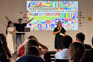 Junge Frau am Mikrofon, hinter ihr sind viele unterschiedliche Länderflaggen an die Wand projiziert.