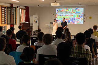 Zwei Jugendliche am Mikrofon, hinter ihr sind viele unterschiedliche Länderflaggen an die Wand projiziert.
