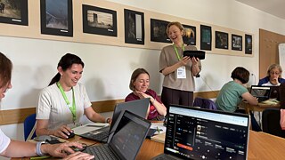 5 Teilnehmende arbeiten mit Laptops an einem großen Tisch, während die Seminarleiterin mit einem tragbaren Lautsprecher in den Händen die gemeinsame Arbeit der Gruppe begleitet.
