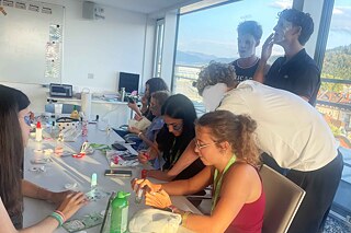 Jugendliche sind um einen Tisch versammelt, auf dem Kosmetika liegen. Sie tragen Masken auf.