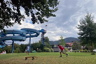 Jugendliche in einem Freibad auf der Wiese, im Hintergrund sieht man eine große Wasserrutsche.
