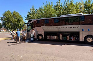 Ein großer Reisebus, davor stehen junge Menschen. Die Gepäckfächer sind voller Koffer.