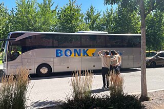 Ein großer Reisebus im Hintergrund, im Vordergrund eine kleine Gruppe junger Menschen, die das Vorgehen beobachten.