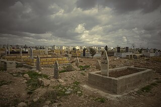 Kurdský hřbitov v Kámišlí u hranice s Tureckem. V dálce je vidět Nusaybin, kurdské město, které se nachází již na turecké straně. Dříve byla obě města propojena a lidé mezi nimi mohli volně přecházet, dnes je odděluje vysoká betonová zeď a hraniční přechody jsou uzavřeny.