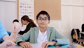 Osoba sedí u stolu v učebně během výuky, zatímco v pozadí probíhá skupinová práce dalších účastníků.