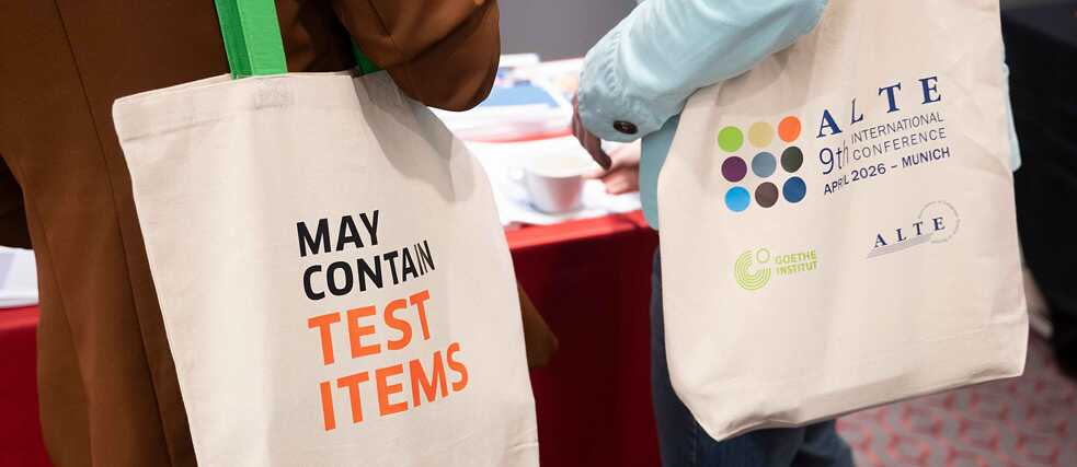 Zwei Konferenzteilnehmende tragen Jutebeutel mit dem Logo der ALTE-Konferenz und der Aufschrift „May contain test items“.
