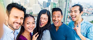 Eine Gruppe Männer und Frauen steht auf einer Dachterrasse. Sie machen ein Selfie. Sie lachen in die Kamera. Eine Person zeigt als Handgeste das Victory-Zeichen. Im Hintergrund ist eine Stadtansicht mit Hochhäusern zu sehen.