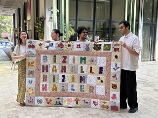 3 Menschen halten lächelnd ein Quilt Banner hoch.