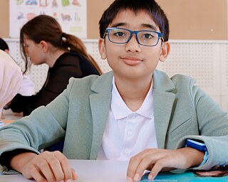 Osoba sedí u stolu v učebně během výuky, zatímco v pozadí probíhá skupinová práce dalších účastníků. 