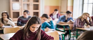 Lernende sitzen in einem Klassenraum und arbeiten konzentriert an schriftlichen Aufgaben. Im Vordergrund befindet sich eine Person an einem Schultisch, während weitere Kursteilnehmende im Hintergrund selbstständig in einem hellen Unterrichtsraum lernen.