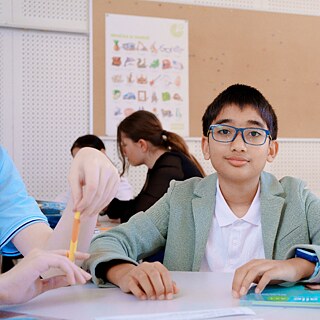 Eine Person sitzt in einem Unterrichtsraum an einem Tisch, während andere Kursteilnehmende im Hintergrund in Gruppen zusammenarbeiten.