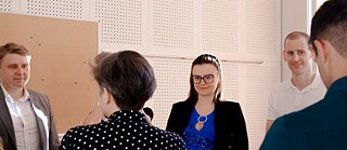 Eine Gruppe von Kursteilnehmenden steht in einem Unterrichtsraum während einer interaktiven Sprachlernaktivität. Mehrere Personen sind in einem Kreis angeordnet und kommunizieren miteinander. Der Unterricht findet in einem modernen Klassenraum mit Pinnwand und schallabsorbierenden Wandpaneelen statt.