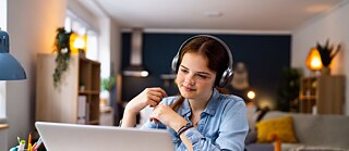 Eine junge Kursteilnehmerin nimmt zu Hause an einem Online-Deutschkurs teil und verfolgt den Unterricht mit Kopfhörern vor einem Laptop. Die moderne und angenehme Lernumgebung ermöglicht konzentriertes Deutschlernen von überall aus.