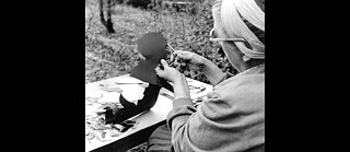 Schwarz-weißes Foto einer Person an einem Tisch im Freien, die mit einer Schere eine Scherenschnittfigur aus schwarzem Papier bearbeitet. Auf dem Tisch liegen weitere ausgeschnittene Papierformen und Arbeitsmaterialien