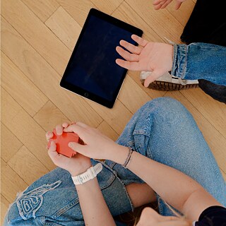 Zwei Personen sitzen auf einem Holzfußboden und arbeiten mit Lernmaterialien. Neben einem Tablet hält eine Person eine rote Karte in der Hand, während eine andere Person auf das Tablet zeigt.
