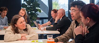 People collaborating and talking around tables in a bright workshop or meeting room.