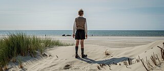 Ein altmodisch angezogenes Kind steht zwischen den Dünen am Strand und sieht aufs Meer hinaus