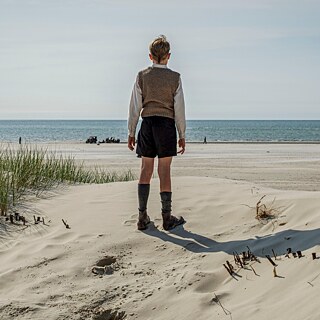 Ein altmodisch angezogenes Kind steht zwischen den Dünen am Strand und sieht aufs Meer hinaus