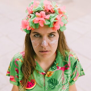 Bild aus der Produktion Guiri. Person mit Blumenkranz und weißer Gesichtsbemalung blickt direkt in die Kamera, gekleidet in ein farbenfrohes, tropisch gemustertes Oberteil