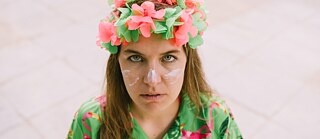 Bild aus der Produktion Guiri. Person mit Blumenkranz und weißer Gesichtsbemalung blickt direkt in die Kamera, gekleidet in ein farbenfrohes, tropisch gemustertes Oberteil