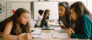 Mehrere junge Schülerinnen sitzen im Klassenzimmer an einem Tisch und besprechen die Aufgaben.