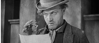 Schwarzweiß Filmstill, das den Kopf eines Manns zeigt, der überrascht auf ein Stück Papier guckt