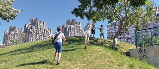 Četvoro dece trči uzbrdo po travi. U pozadini se vide novobeogradski blokovi.