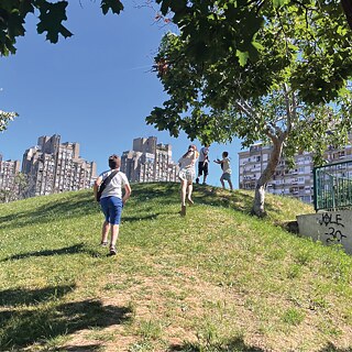 Četvoro dece trči uzbrdo po travi. U pozadini se vide novobeogradski blokovi.