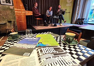Podiumsgespräch auf einer Bühne, davor Tisch mit Zeitschriften. 