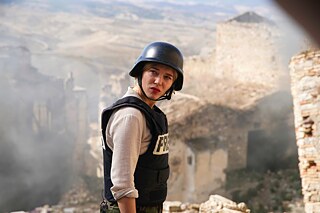 Eine Frau mit Schutzhelm und kugelsicherer Weste mit der Aufschrift „PRESS“ vor einem Kriegsschauplatz.