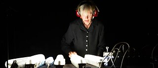 Die deutsche Klangkünstlerin Christina Kubisch steht in einem abgedunkelten Raum vor einer Installation mit leuchtenden elektronischen Geräten und Kabeln. Mit speziellen Kopfhörern untersucht sie die Klangwelten elektromagnetischer Felder, die für ihr Projekt „Electrical Walks“ charakteristisch sind.