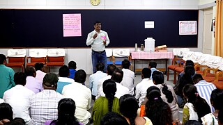 One person standing against a blackboard talking to a group of sitting people, with their backs to the camera