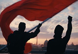 Zwei schwarze Figuren im Gegenlicht schwenken eine rote Flagge; im Hintergrund die Skyline einer Stadt bei Sonnenuntergang.