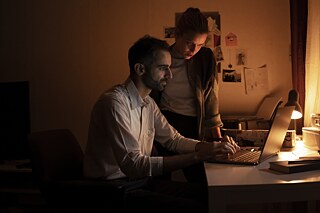 Ein Mann und eine Frau an einem schwach belichteten Schreibtisch mit PC.