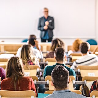 Rear view of a group of university students seated in a lecture hall, with a lecturer standing at the front, softly blurred and out of focus.