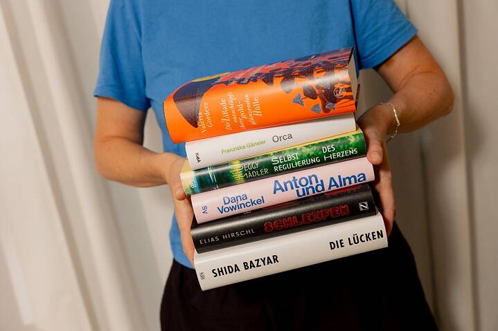 Eine Person in einem blauen T-Shirt hält vor hellem Hintergrund einen Stapel von sechs Büchern. Zu erkennen sind die Buchcover und Buchrücken der nominierten Titel der Shortlist des Deutschen Buchpreises. Die Bücher sind übereinandergestapelt und werden mit beiden Händen getragen.