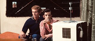 Two people sit closely together on a sofa, watching television; one drinks from a glass bottle.