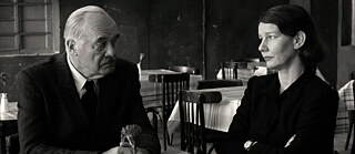Black-and-white photograph of two people seated opposite each other at a table in a sparsely furnished restaurant.