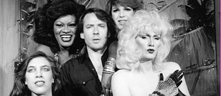 Black-and-white photograph of five people posing closely together in an ornate nightclub interior, wearing theatrical clothing, hairstyles and accessories. The image is framed by a thick, rounded bright pink border.