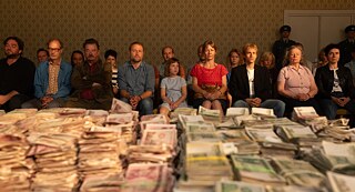 escena de la película zwei zu eins. Grupo de personas sentadas frente a una mesa que contiene pilas de dinero en efectivo
