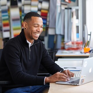 A young man sits at a desk smiling and moving the mouse. He looks at a screen.