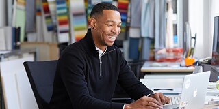A young man sits at a desk smiling and moving the mouse. He looks at a screen.